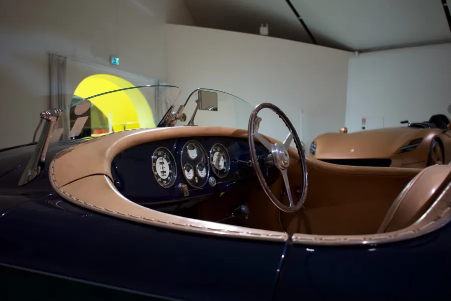 A vintage open two-seater in a museum, its wooden steering wheel and round gauges framed by a low curved windshield