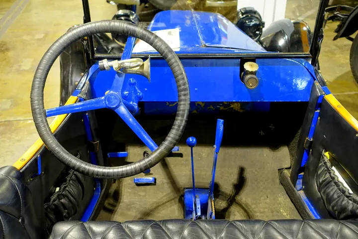 Cockpit of a 1914 roadster: a large black steering wheel, exposed floor levers and a blue-painted body with no doors or roof