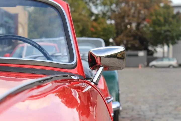 Close-up of a red classic convertible’s chrome side mirror and windshield frame, with other vintage cars parked behind it