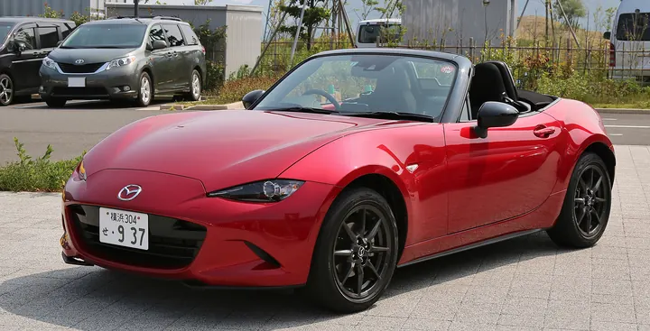 A red modern two-seat roadster with its soft top folded, parked on a paved lot