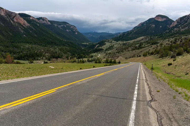 A two-lane highway with a yellow center line leading toward rugged mountains under a cloudy sky on the Beartooth Highway